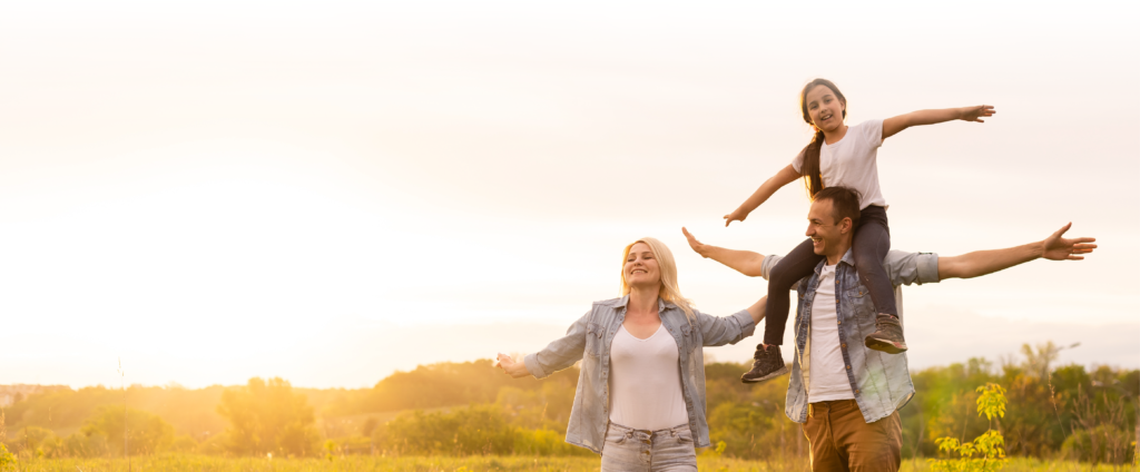 Man and woman smiling with their arms out in a field. The sun is setting in the distance and a little girl sits on the man's shoulders