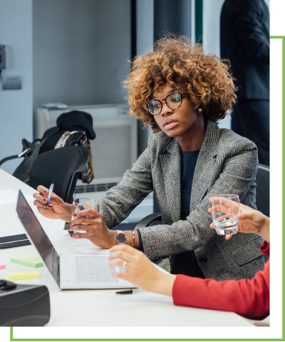 A woman looks at a laptop in an office setting. Another person sits next to her holding a glass.