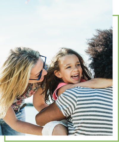 A little girl holds onto an adult around their neck, a woman wearing sunglasses smiles next to them